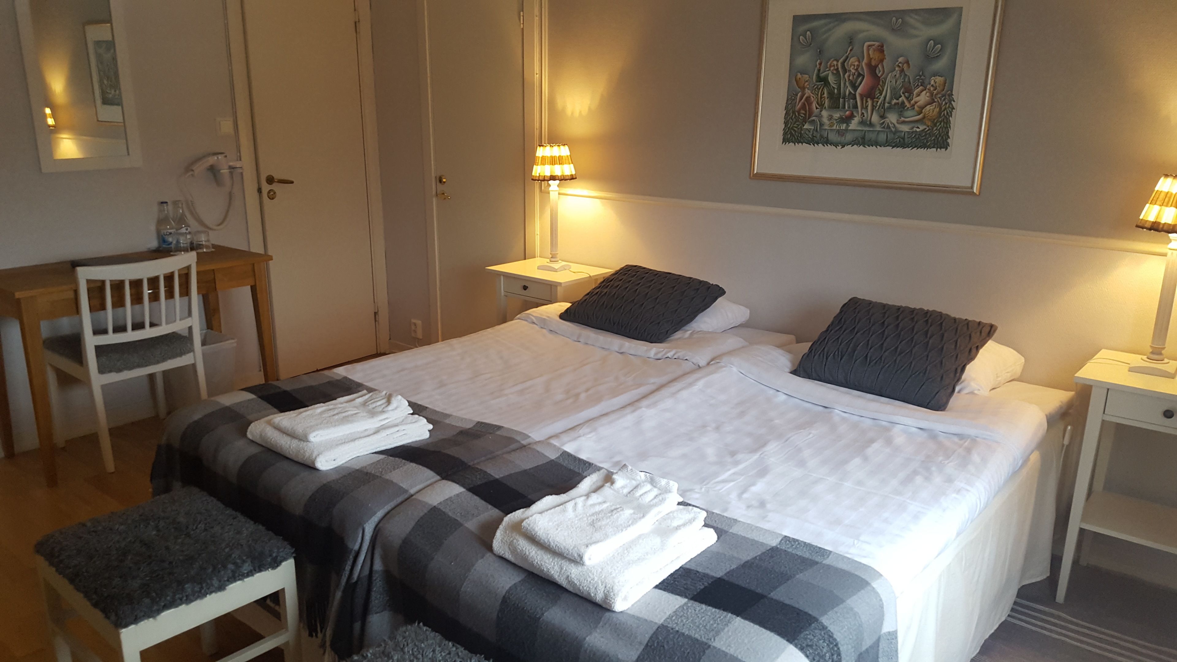 Una cama bien hecha con un edredón de cuadros, dos lámparas de noche, una silla y un escritorio con lámpara.