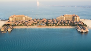 Luftaufnahme eines Küstenresorts mit mehreren Gebäuden, einem Strand und einer segelartigen Konstruktion in der Ferne.