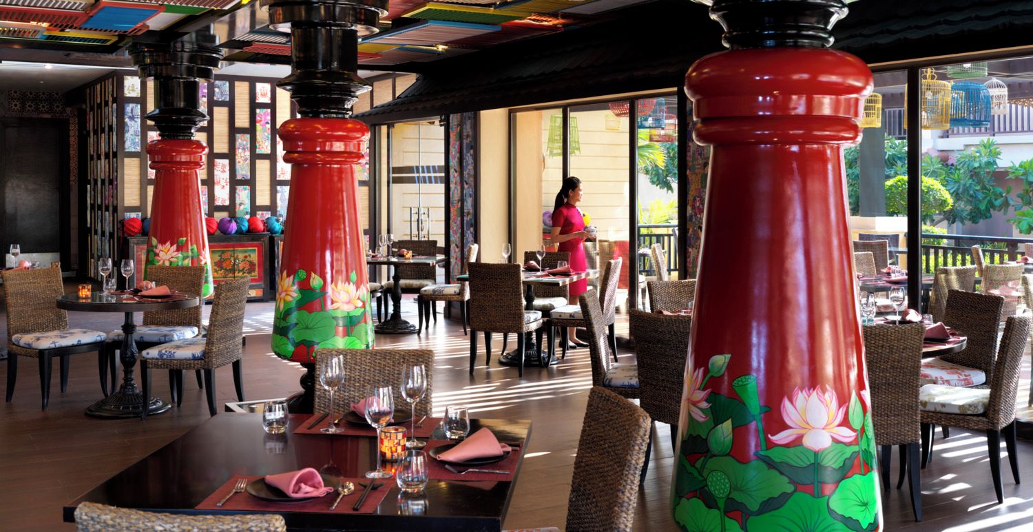 A restaurant with wicker furniture, red pillars with lotus designs, and a view of the outdoors.