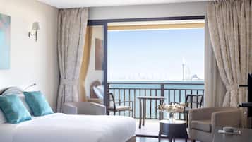 Habitación de hotel con una cama grande, dos sillones, una mesita con un jarrón de flores y un balcón con vistas a la ciudad y un puente.
