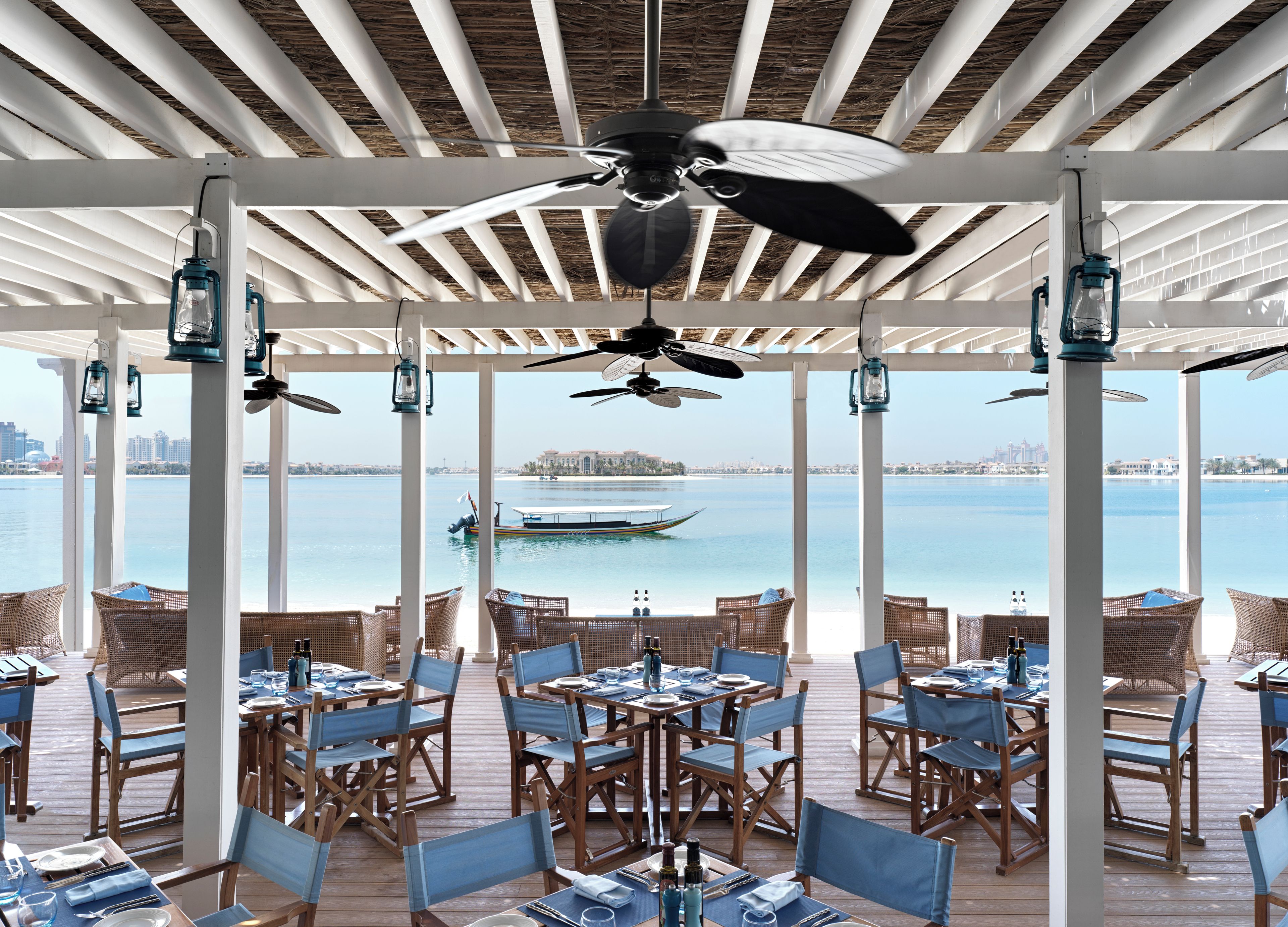 An outdoor dining area with tables and chairs, a ceiling fan, and a view of the water and cityscape.
