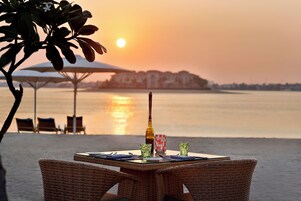 A table set for two with a bottle, glasses, and cutlery, overlooking a calm body of water with a sunset in the background.