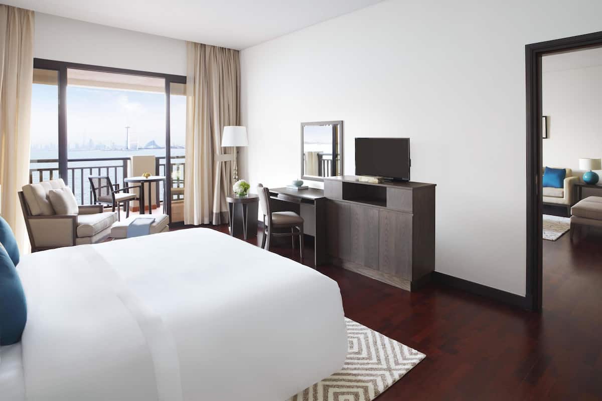 Een hotelkamer met een groot bed, een bureau met een televisie, een balkon met uitzicht op de stadshemel en een zithoek met een bank en stoelen.