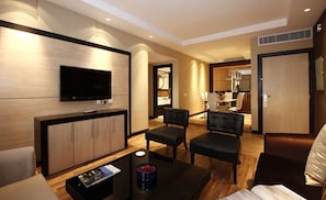 A modern hotel room with a flat-screen TV mounted on the wall, a sofa, two armchairs, a coffee table, and a small side table.