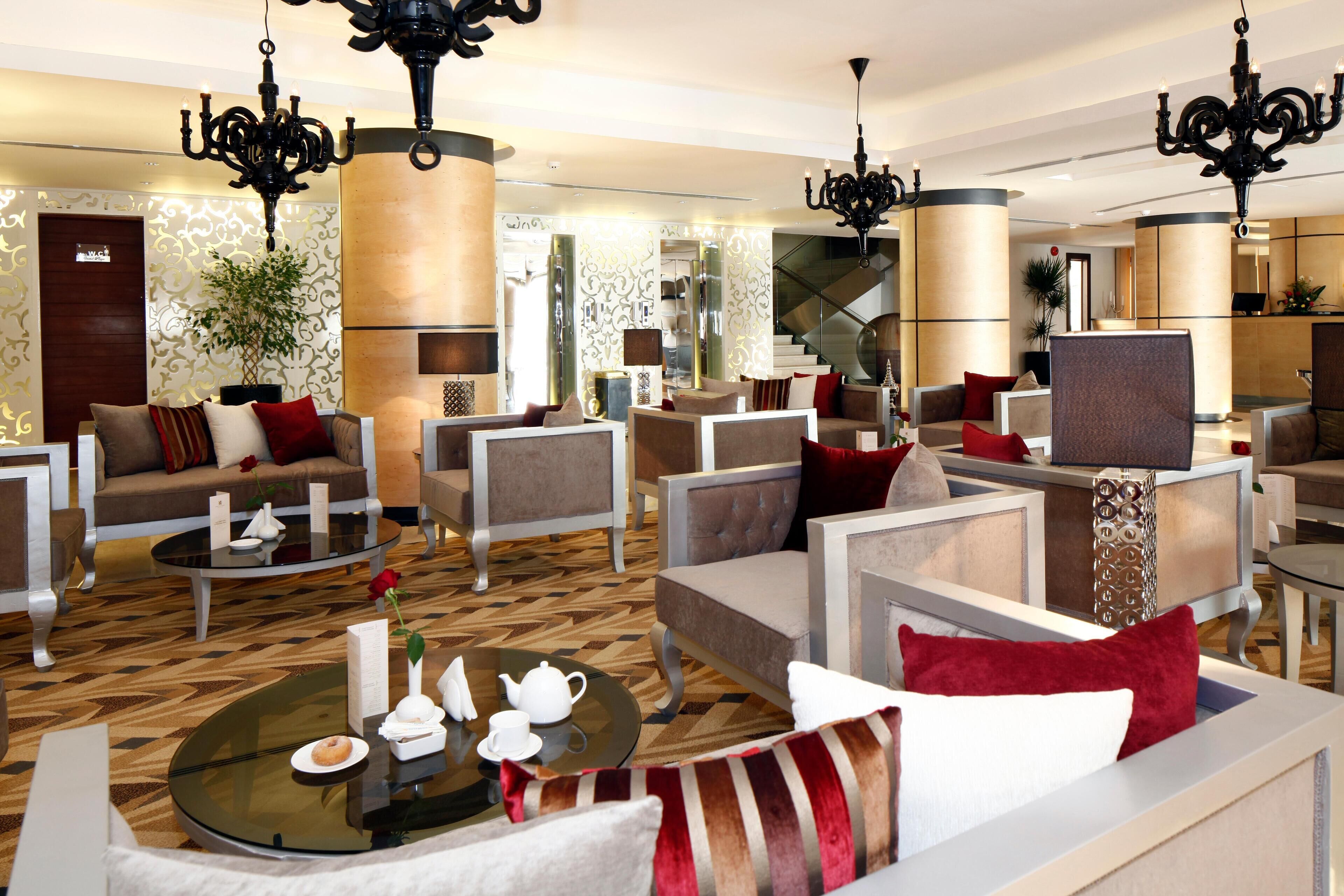 Un moderno vestíbulo de hotel con una variedad de zonas de estar, incluyendo sofás y sillones, dispuestas alrededor de una mesa de centro. Varias arañas cuelgan del techo y se observa una escalera al fondo.