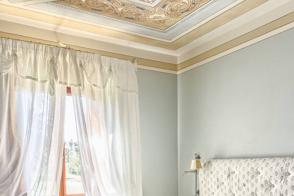 Ein Schlafzimmer mit einem großen Bett, einem Nachttisch, einer Lampe und einem Fenster mit durchsichtigen Vorhängen.