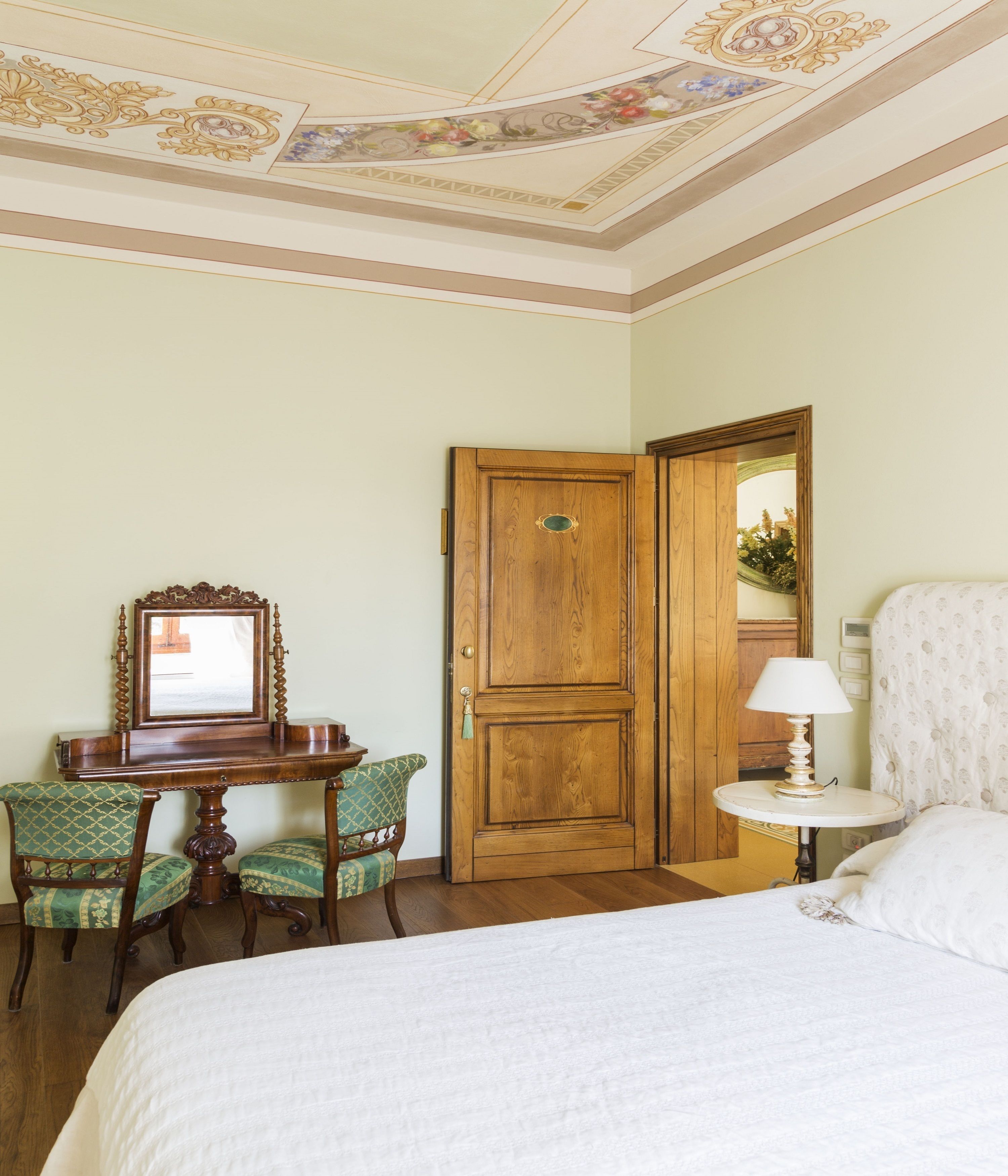 Ein Hotelzimmer mit einem Bett, einer Holztür, einem Schreibtisch mit Spiegel und zwei Polsterstühlen.