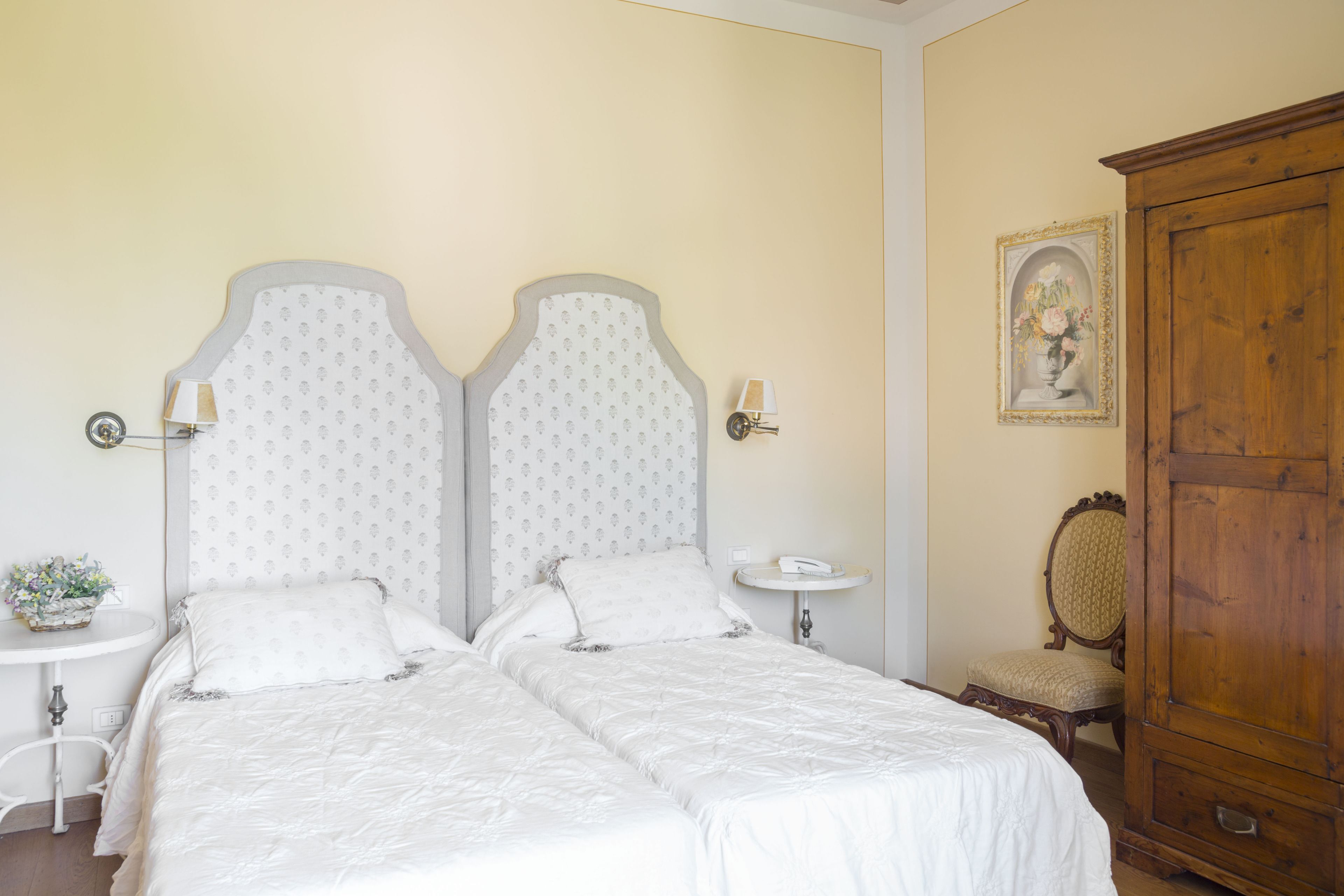 Una cama doble con una colcha blanca y dos almohadas, una mesita redonda con una lámpara, un armario de madera y un cuadro en la pared.