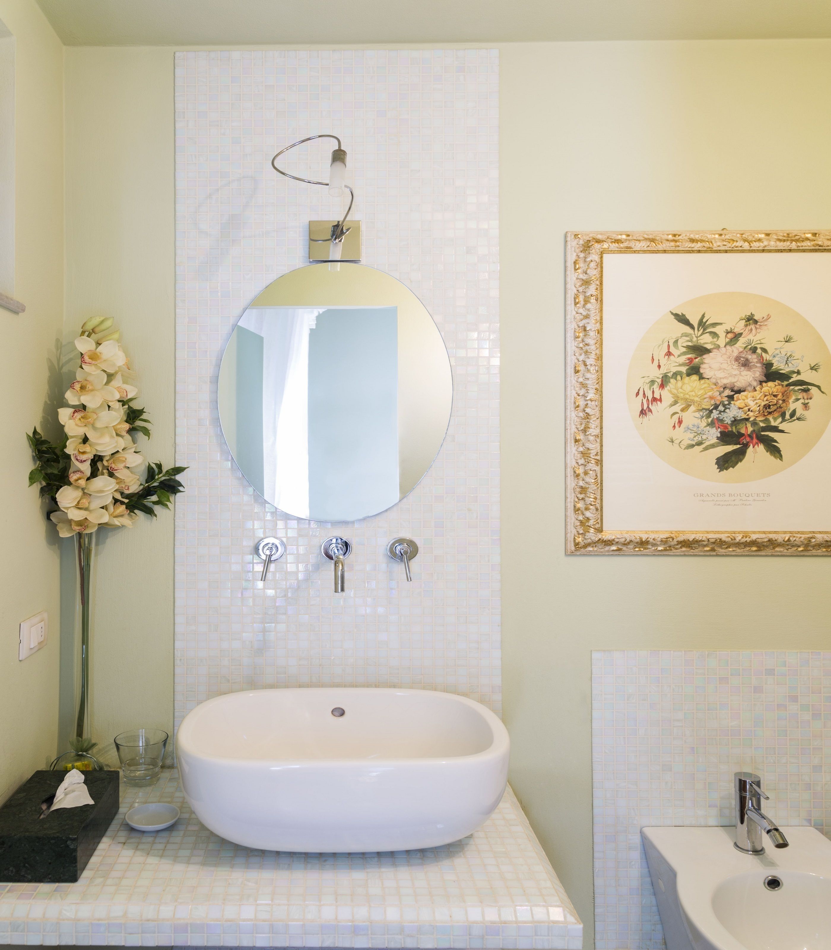 Ein Badezimmer mit einem großen, ovalen Waschbecken, einem Spiegel, einem gerahmten Blumenbild und einer Vase mit Blumen.