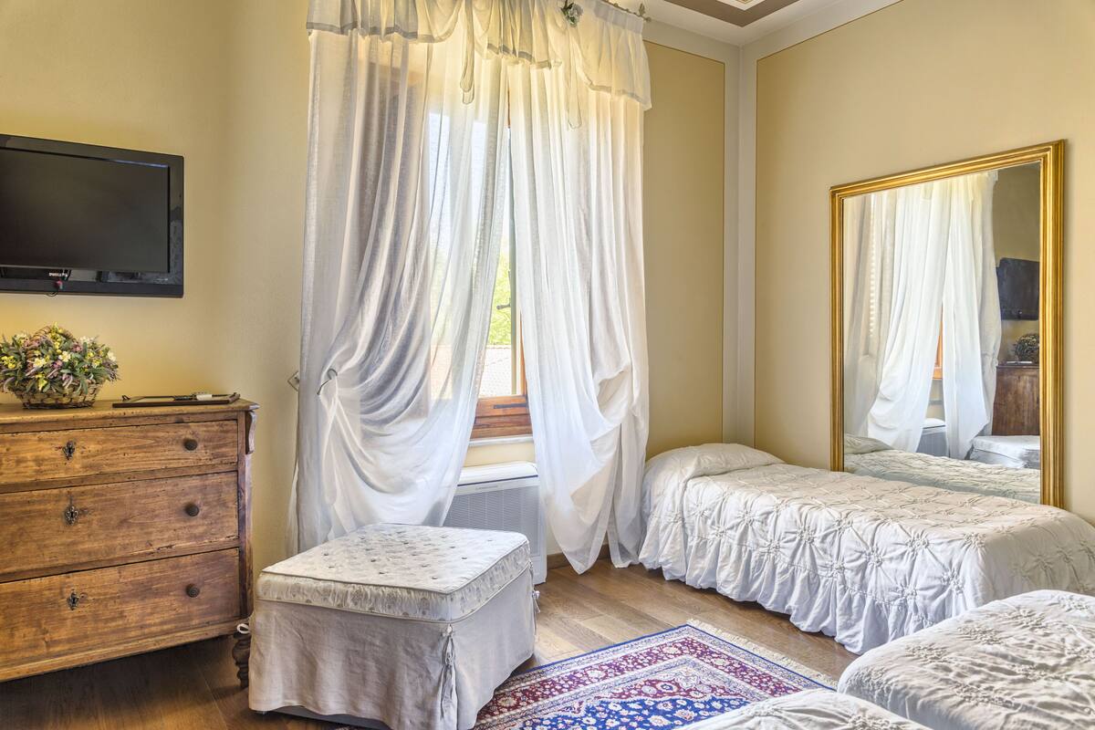 Ein Schlafzimmer mit einem Holzkasten, einem Nachttisch, einem großen Spiegel und einem Fenster mit transparenten Vorhängen.