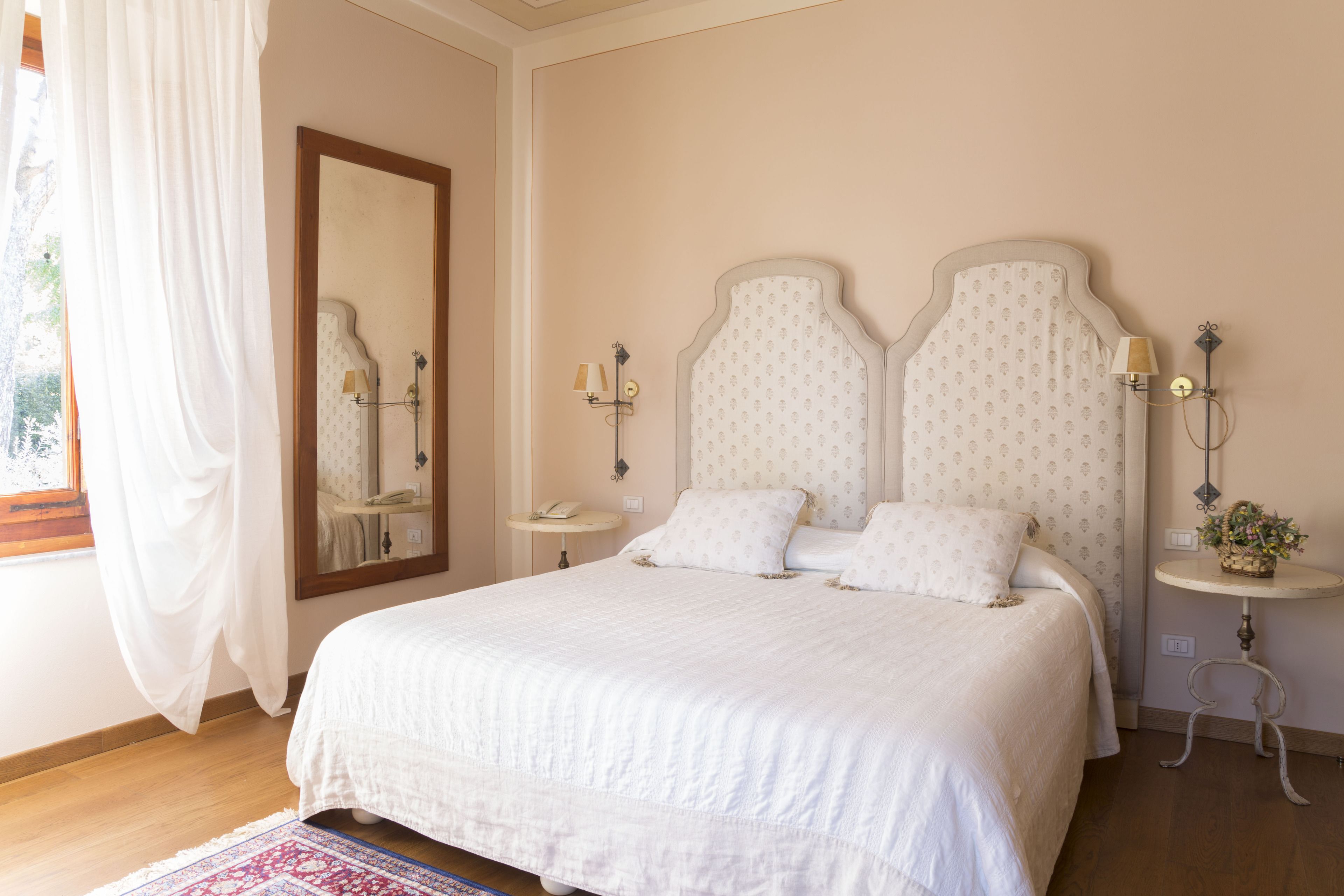 A bedroom with a large bed, a bedside table, a nightstand, a mirror, and a window with curtains.