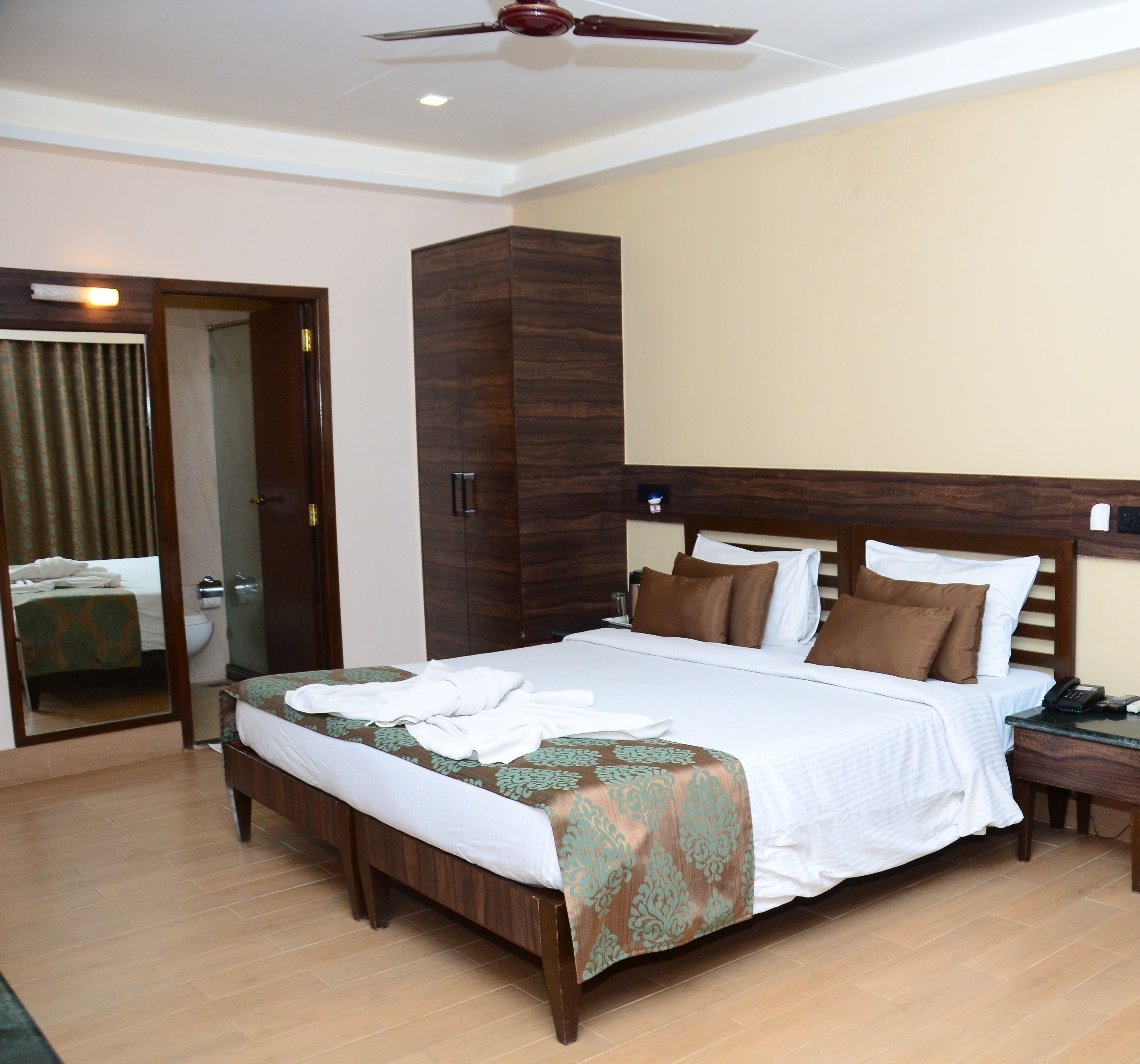 A hotel room with a large bed, a wooden wardrobe, a bedside table with a phone, and a ceiling fan.