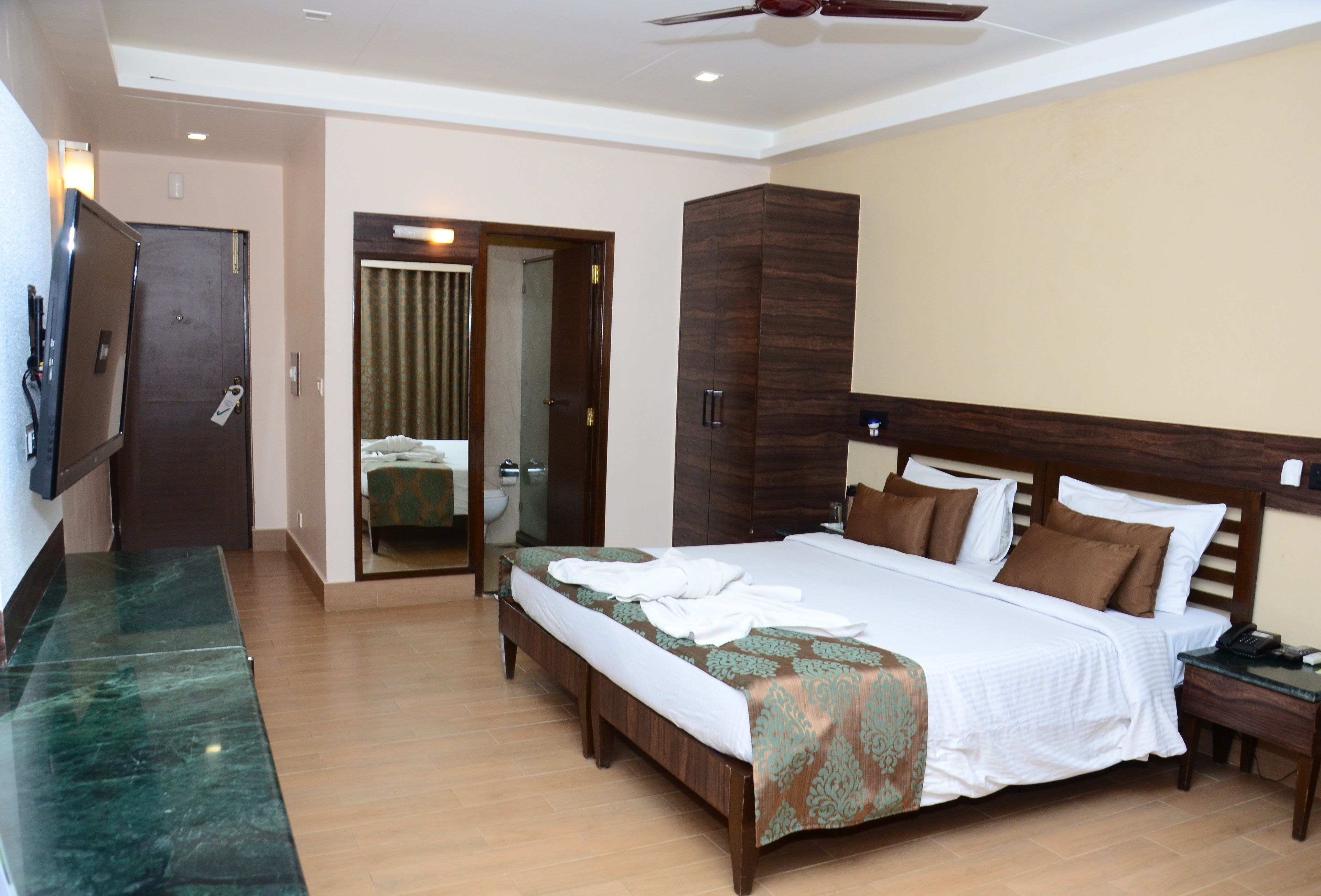 A hotel room with a large bed, a wooden headboard, a nightstand, a desk, a mirror, a wardrobe, and a television.