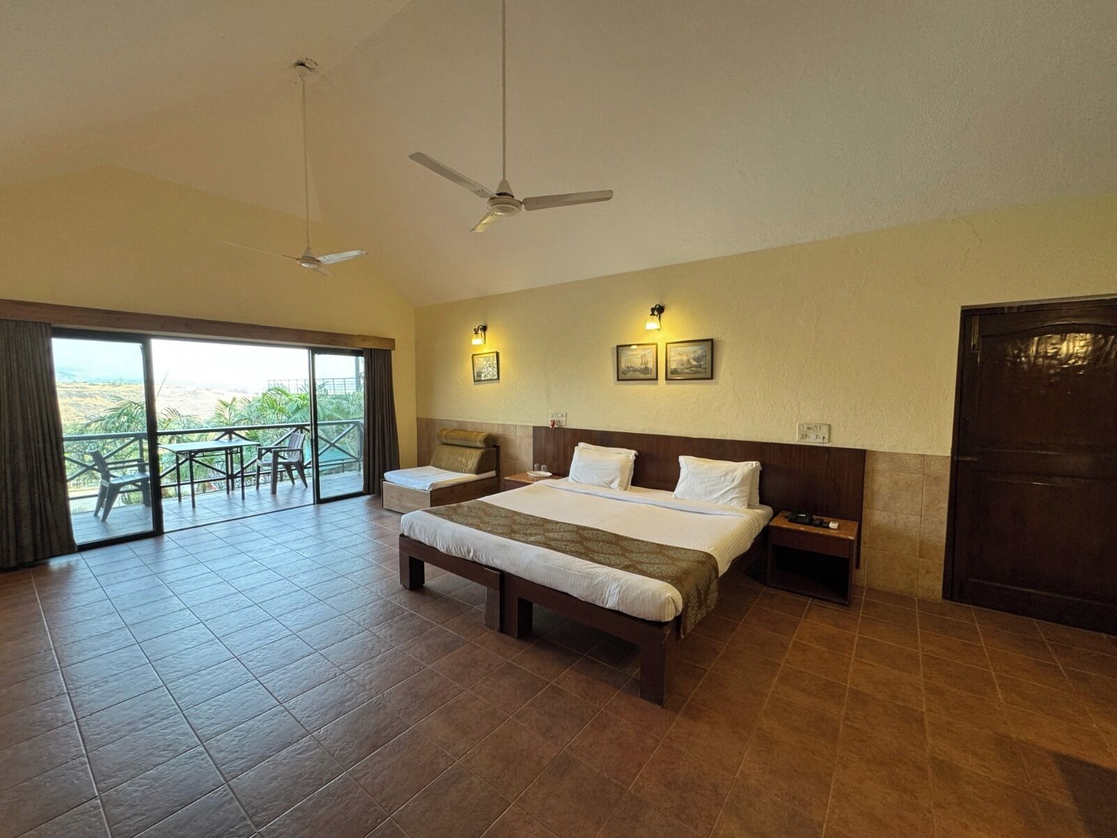 A hotel room with a large bed, two bedside tables, a ceiling fan, and a balcony with outdoor seating.