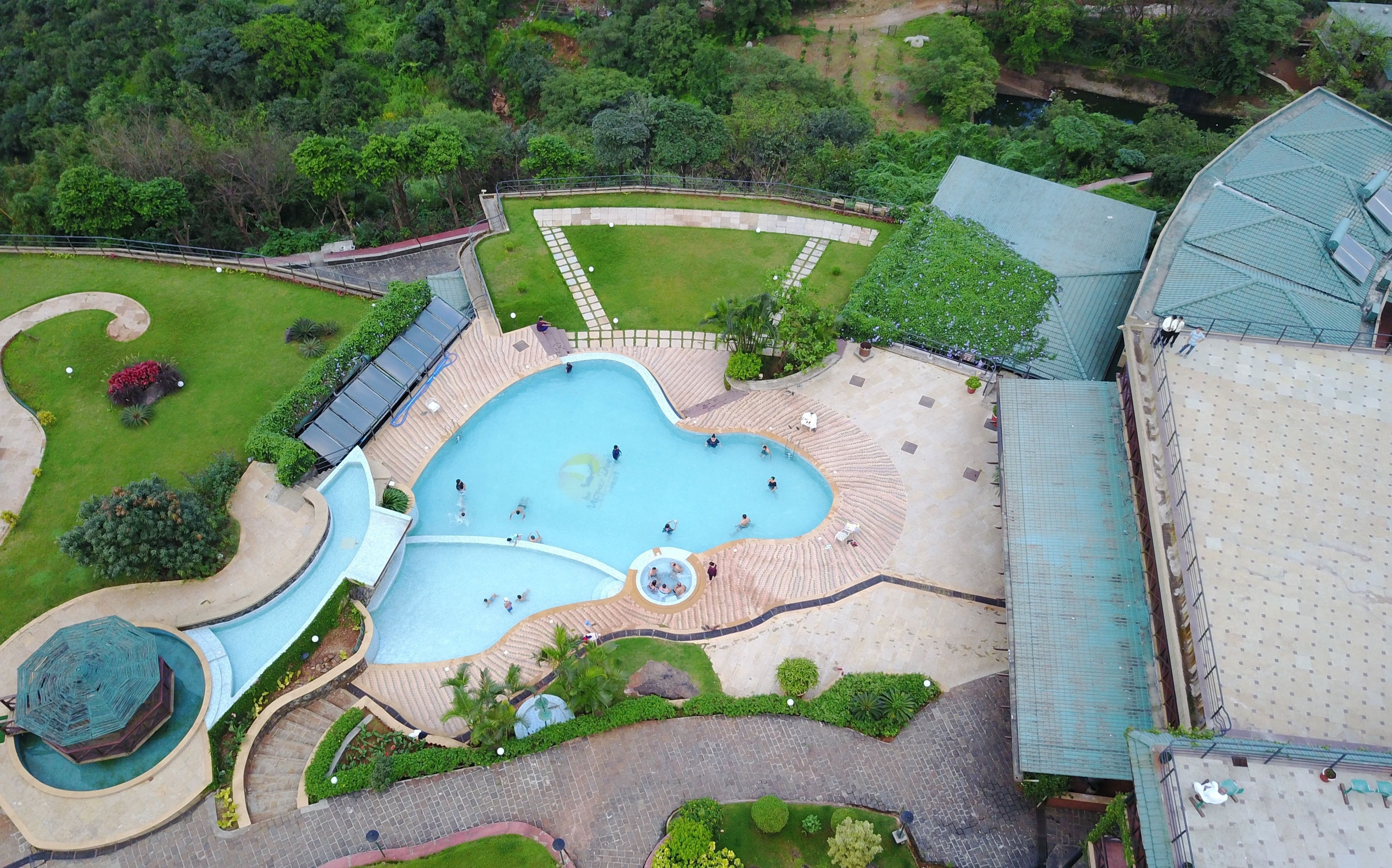 Une vue aérienne d’un hôtel doté d’une grande piscine, de jardins aménagés et d’un espace couvert avec des sièges.