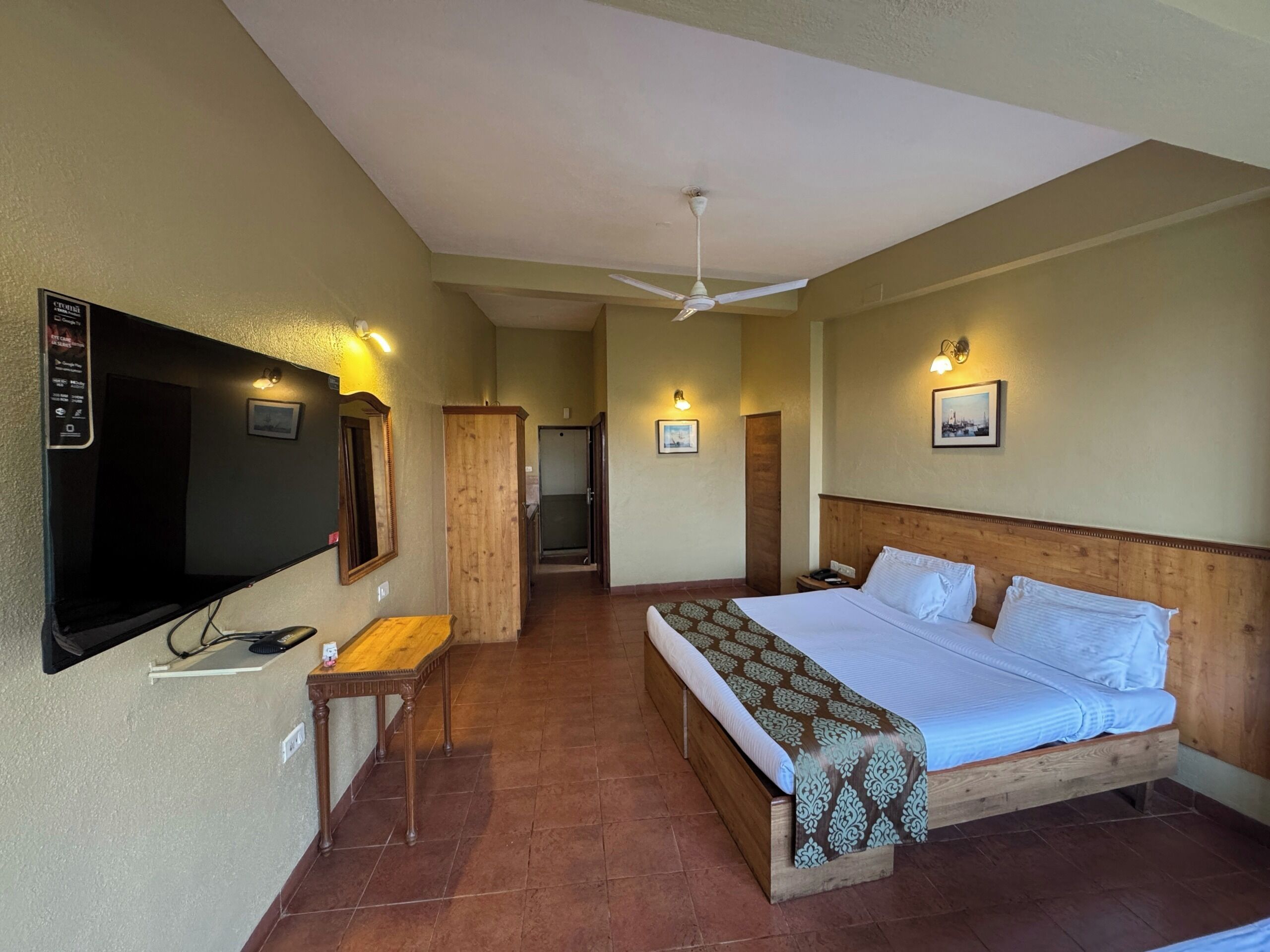 Habitación de hotel con cama, escritorio, televisor y ventilador de techo.
