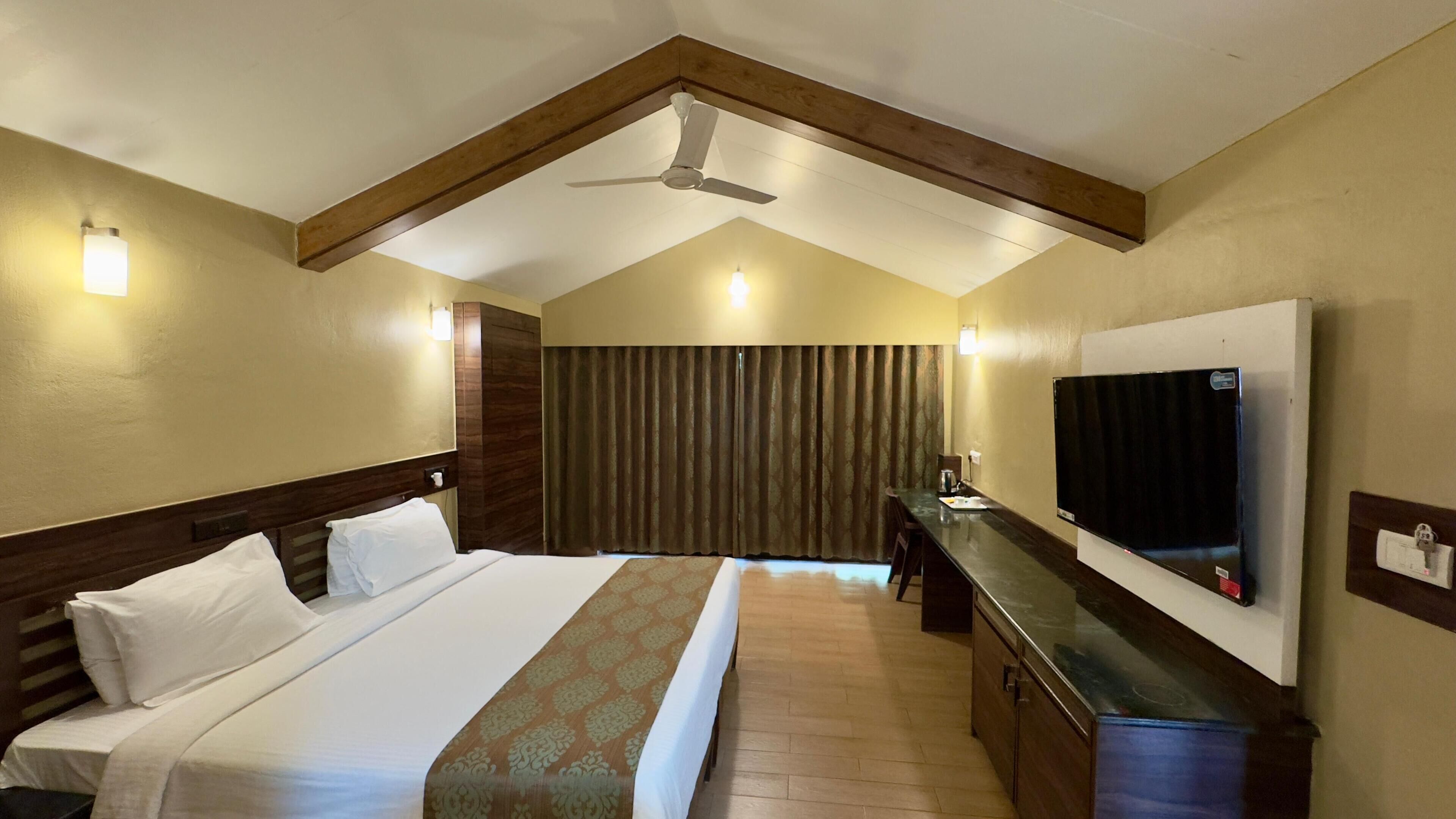 A hotel room with a bed, a desk, a flat-screen TV, a ceiling fan, and a window with curtains.