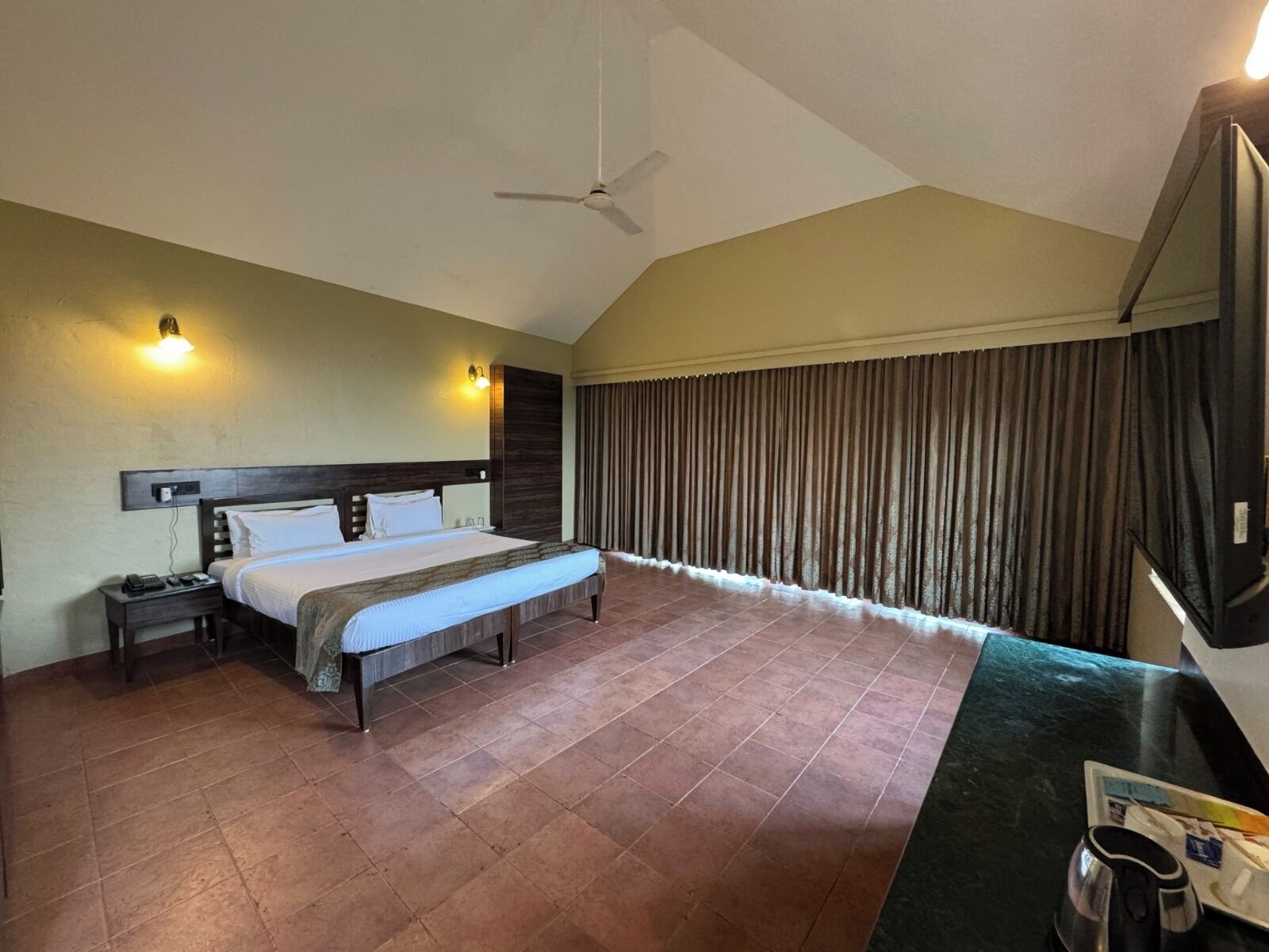 A hotel room with a bed, bedside tables, a ceiling fan, and a view of the outside through curtains.