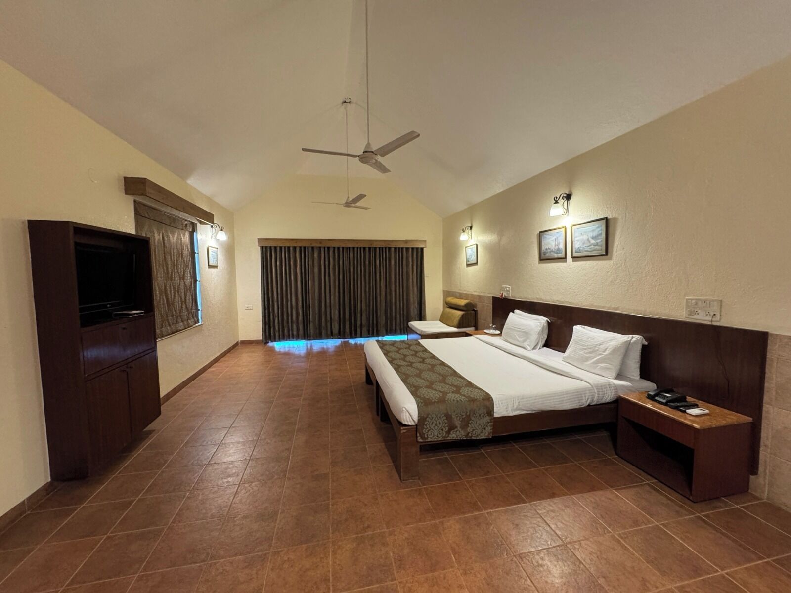 A hotel room with a large bed, a TV, a ceiling fan, and a window with curtains.