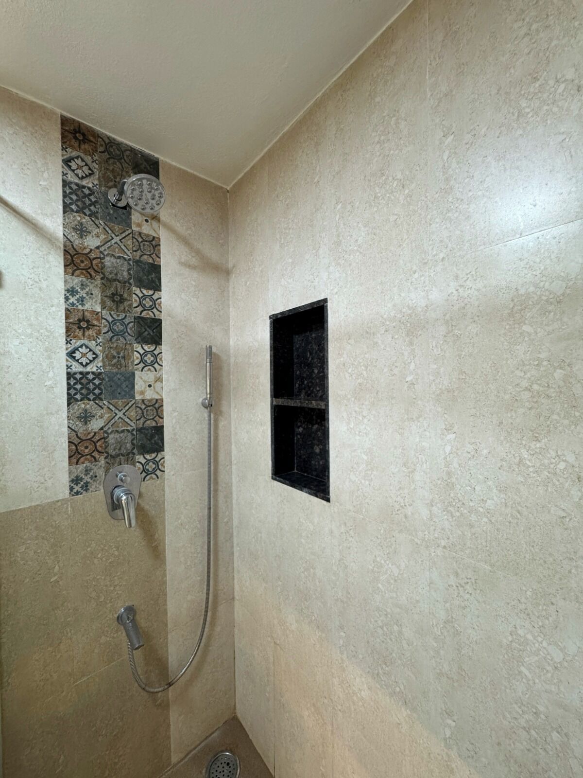 A shower with a hand shower and a wall-mounted faucet, a tiled shower area, and a small black shelf.