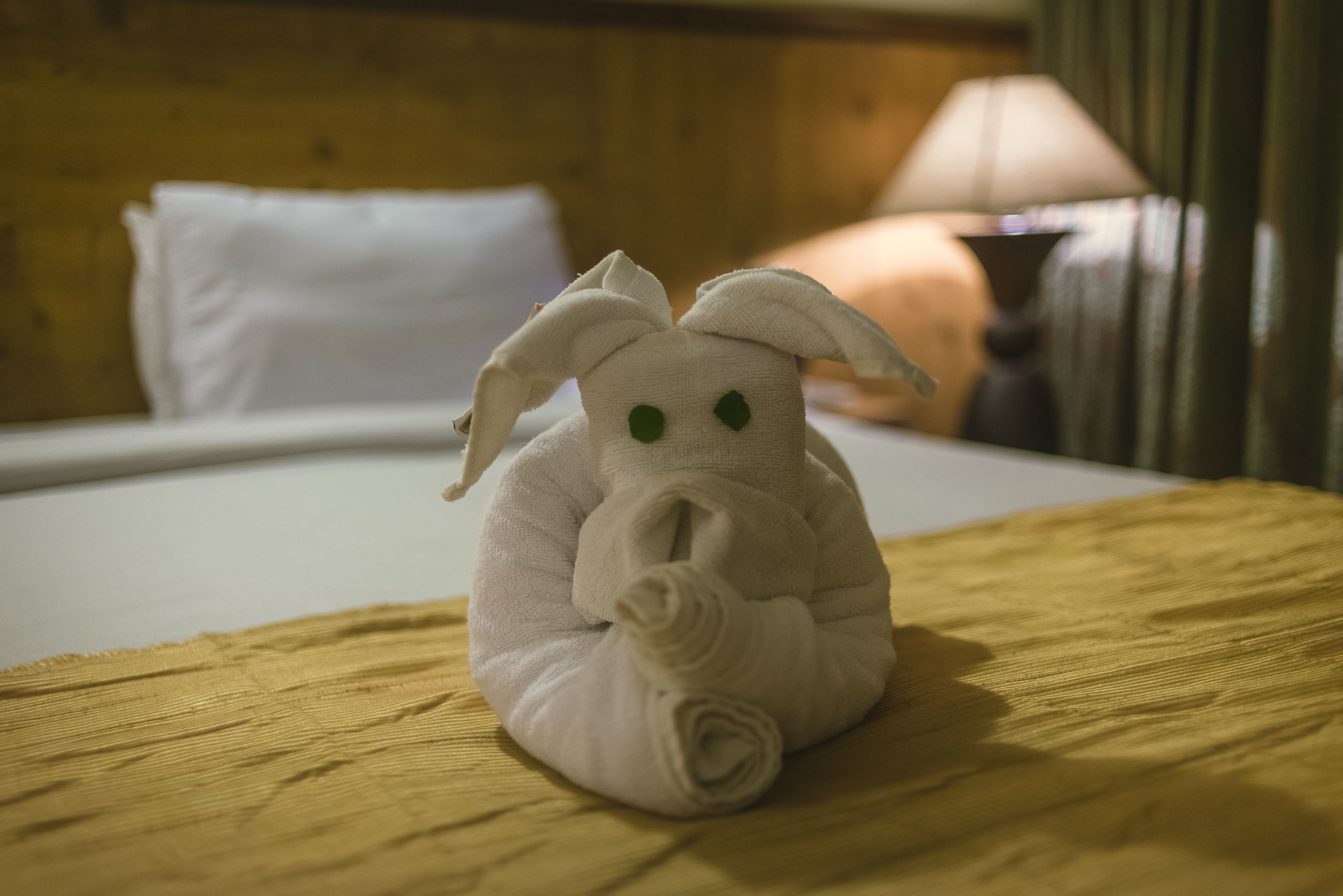 A towel shaped like a bunny on a bed with a wooden headboard and a lamp in the background.
