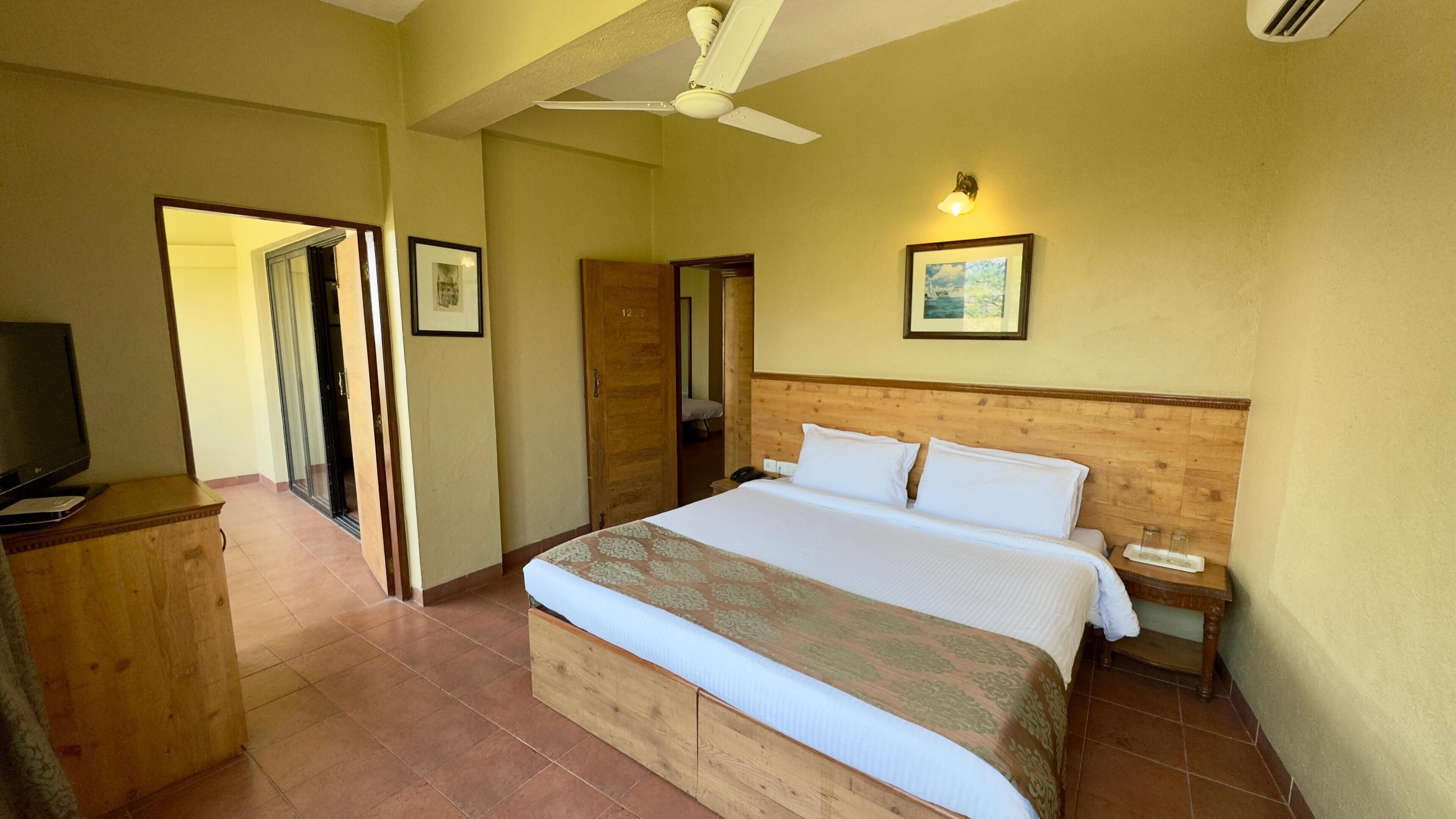 Ein Hotelzimmer mit einem großen Bett, einem Schreibtisch mit Fernseher, einem Deckenventilator und einem gerahmten Bild an der Wand.