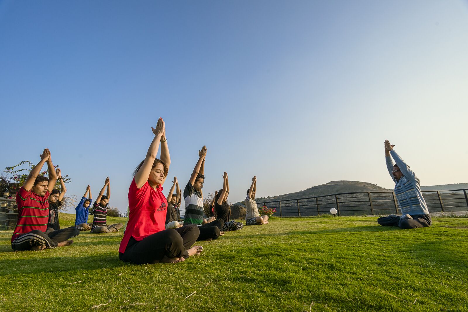 A group of people performing yoga on a grassy field with a clear sky and a hilly landscape in the background.