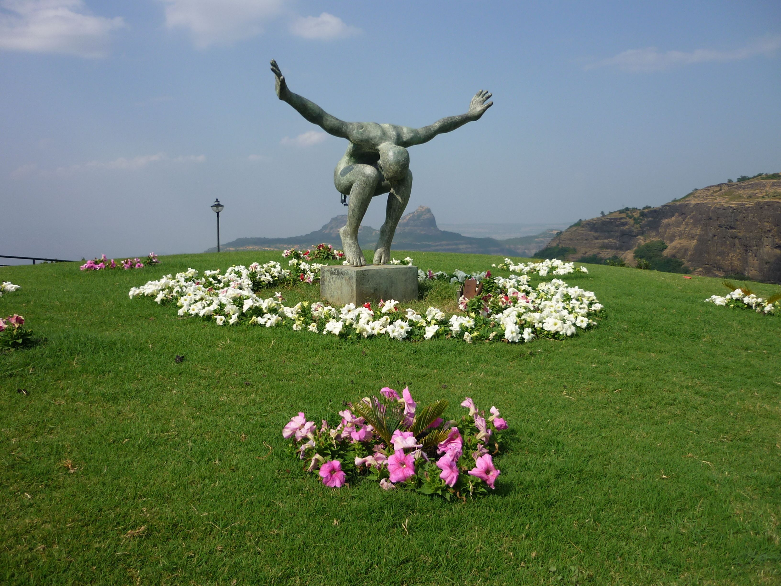 A statue of a person with arms raised, surrounded by flowers on a grassy hill with a mountain in the background.