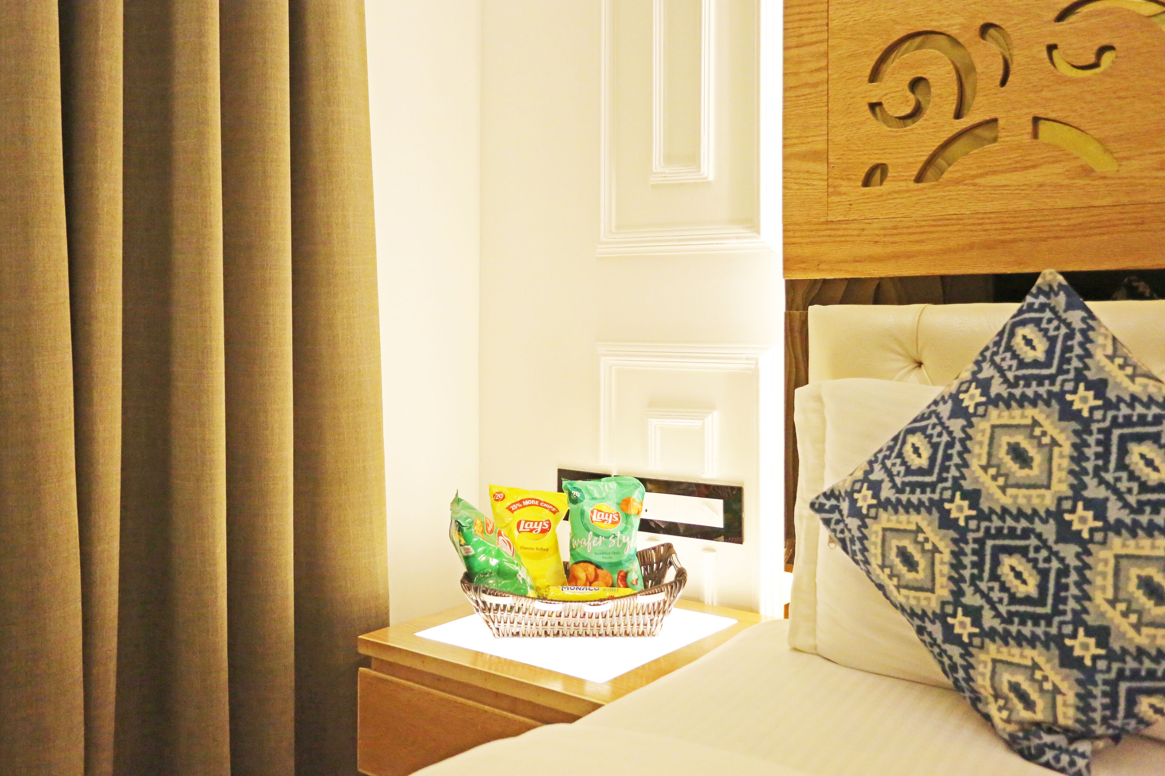 A hotel room with a bed, a bedside table with a basket of snacks, a patterned pillow, and curtains.