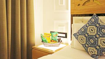 A hotel room with a bed, a bedside table with a basket of snacks, a patterned pillow, and curtains.