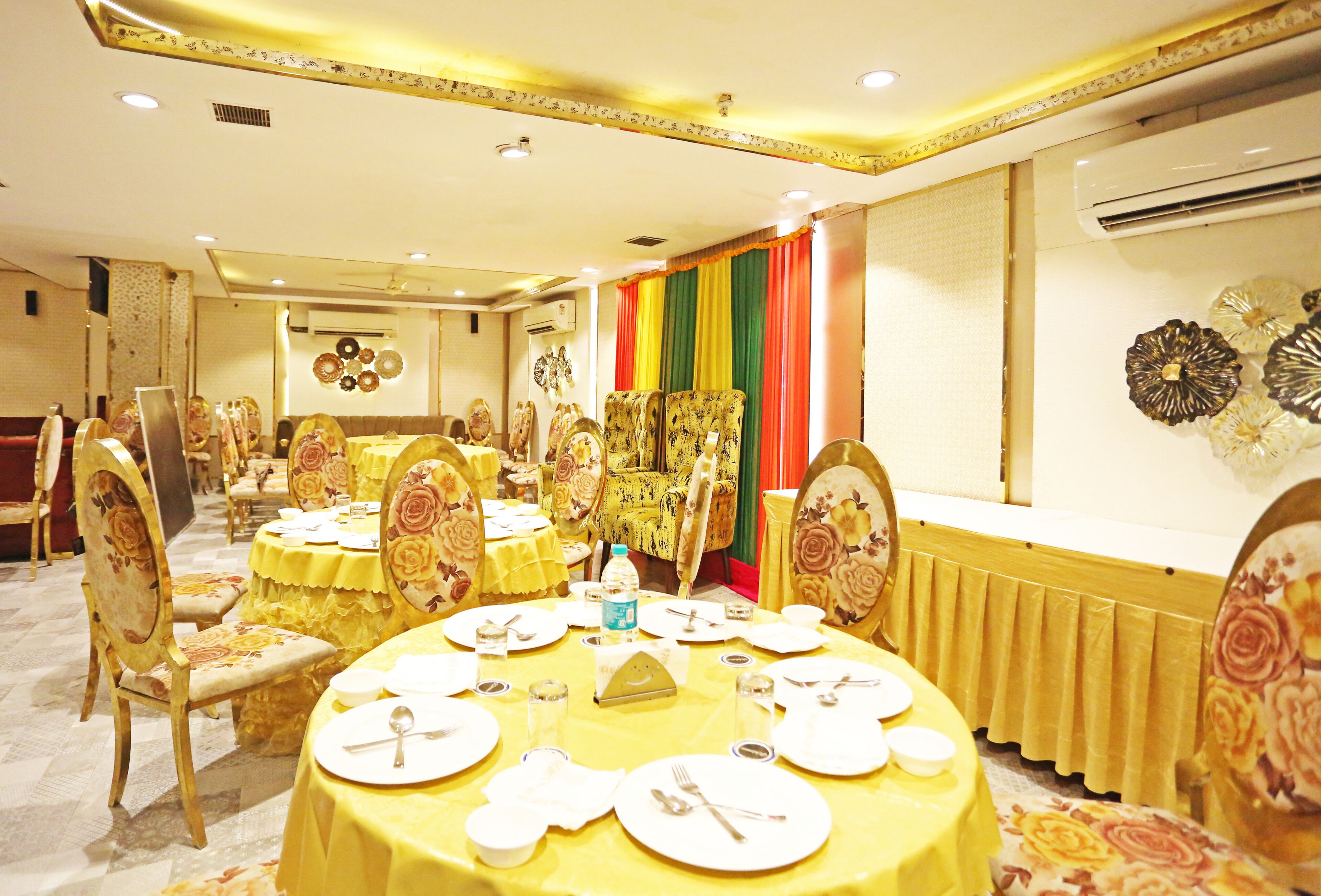 A banquet hall with round tables covered in yellow tablecloths, floral chairs, and a colorful drape in the background.