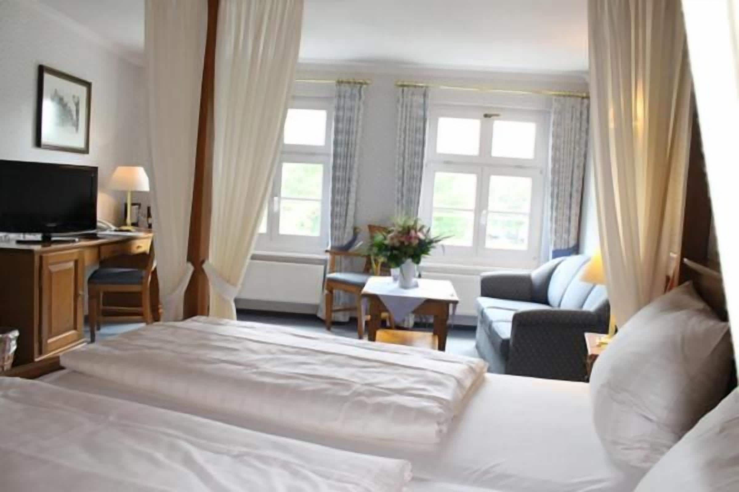 Habitación de hotel con una cama grande, un escritorio con televisor, una silla, una mesita con un florero y un sofá.