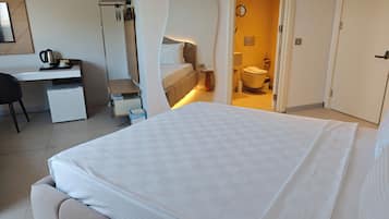 Una habitación de hotel moderna con una cama, un escritorio, una silla, un refrigerador pequeño y un baño visible a través de una puerta abierta.