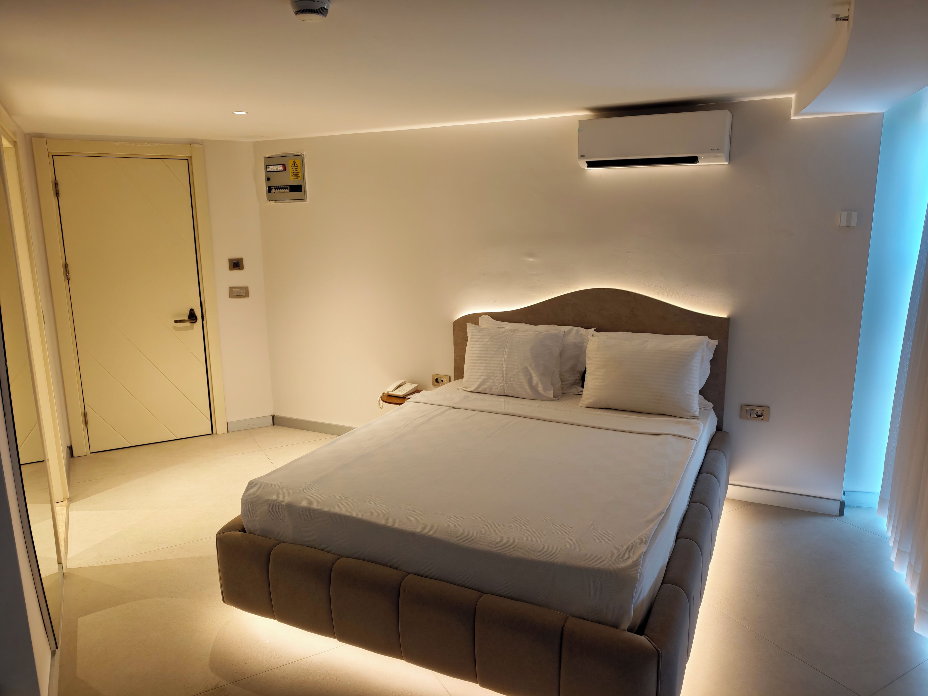 A hotel room with a bed, bedside table, and an air conditioning unit.