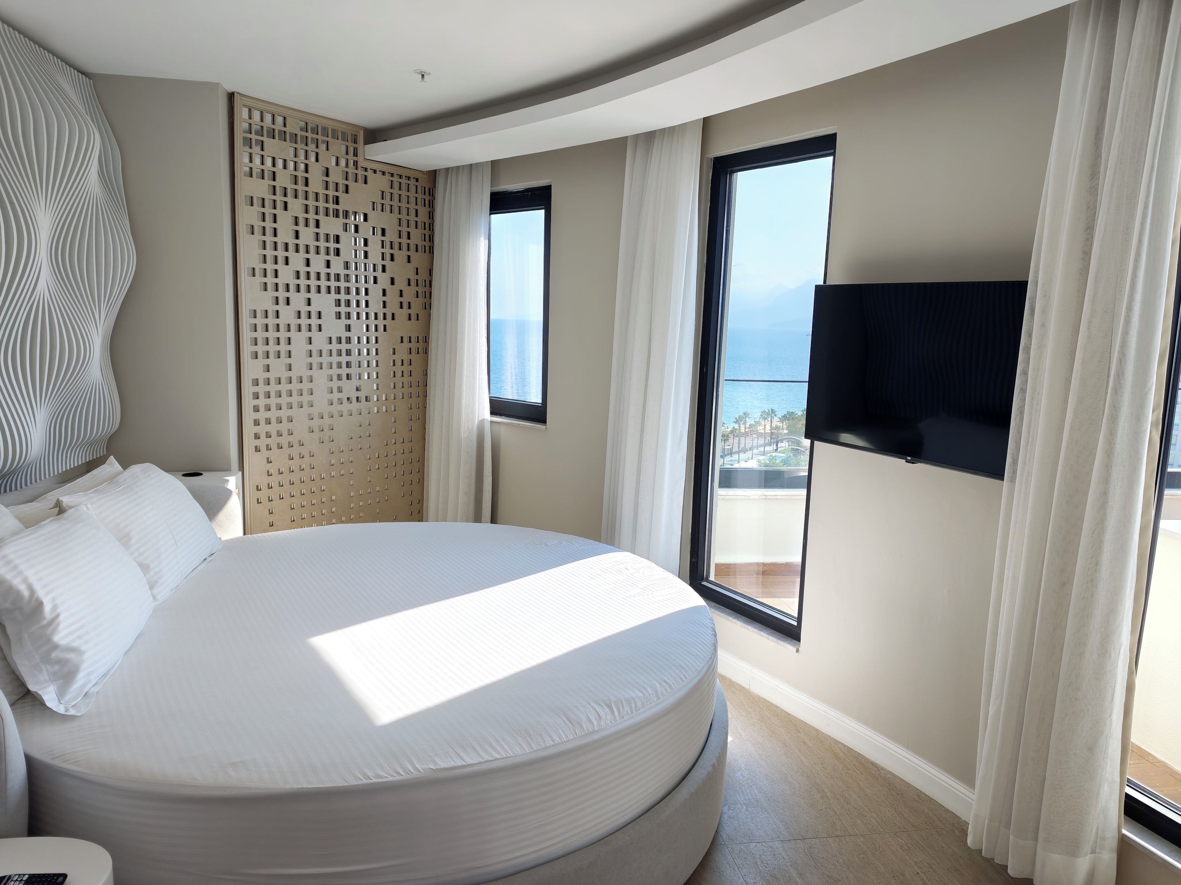 Habitación de hotel con una cama grande, un televisor montado en la pared y vista al mar por la ventana.