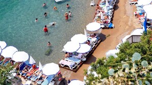 Een kuststrand met helderblauw water, ligbedden, parasols en een stenen muur.