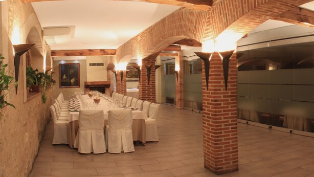Una larga mesa de comedor con manteles blancos y sillas, ubicada en una sala con arcos de ladrillo y divisiones de vidrio modernas.