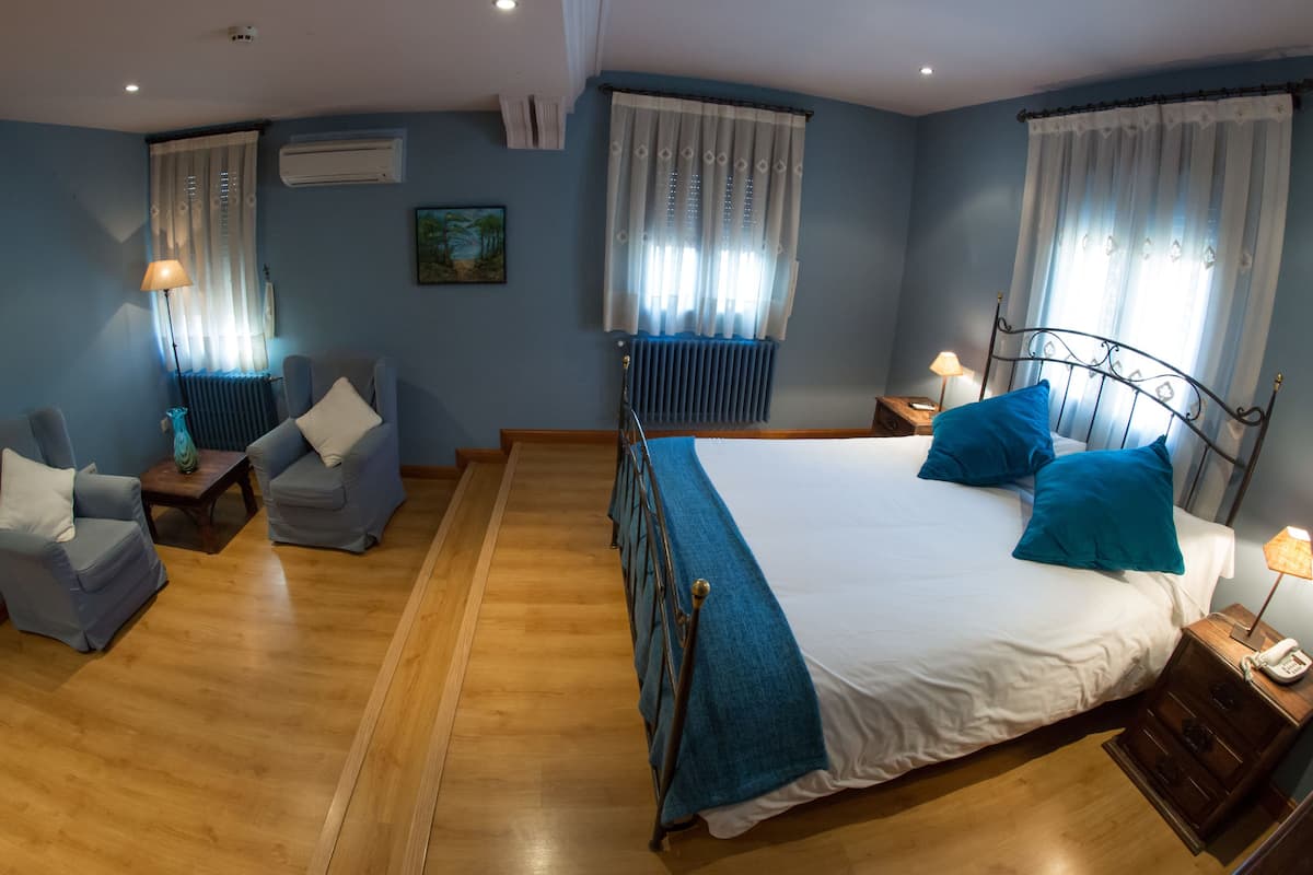 Habitación de hotel con cama, dos sillones, suelo de madera, radiador, un cuadro en la pared y ventana con cortinas.