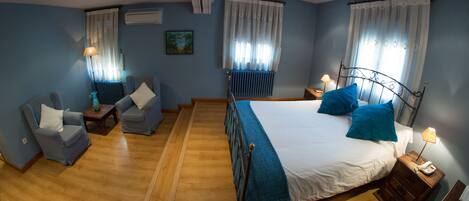 A hotel room with a bed, two armchairs, a wooden floor, a radiator, a painting on the wall, and a window with curtains.