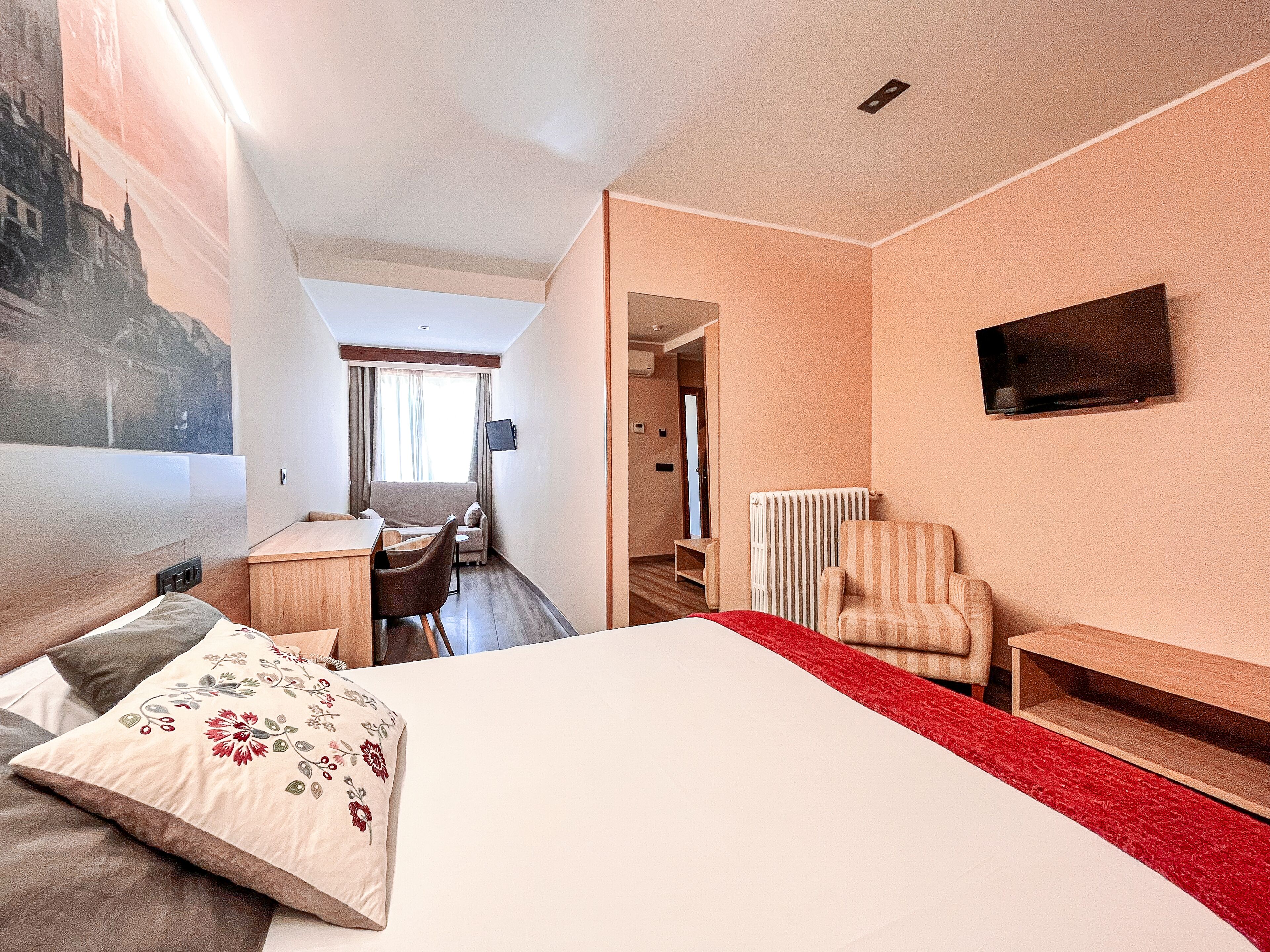 A hotel room with a bed, a desk, a chair, a TV, and a radiator.