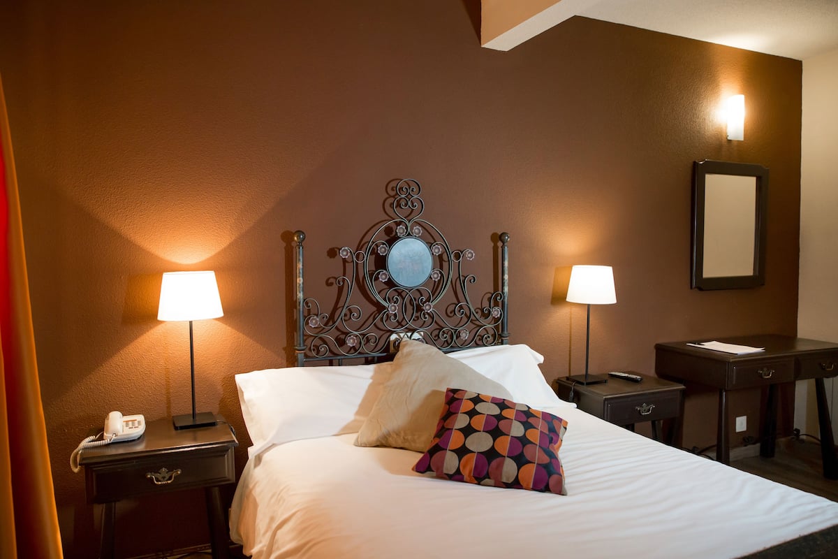 Habitación de hotel con cama, dos lámparas de noche, mesita de noche con teléfono, escritorio con espejo y luz en la pared.