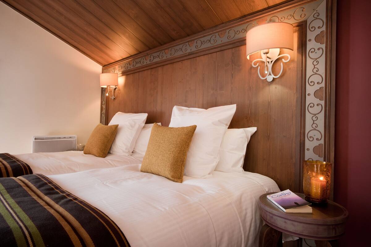 A double bed with white and brown pillows, a bedside table with a book and a candle, and a wall with decorative molding.