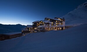 A multi-story hotel with balconies and snow-covered roofs, situated on a snowy slope with mountains in the background. - Le Hameau du Kashmir (Les Belleville)