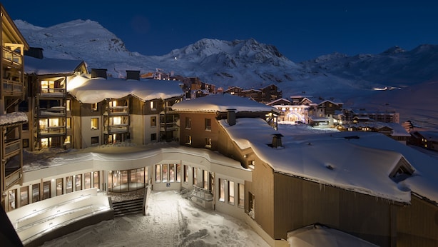 Un complejo turístico cubierto de nieve con varios edificios, un telesilla y montañas al fondo.