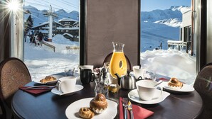 Una mesa de comedor con alimentos para el desayuno y un vaso de jugo de naranja, con vistas a un paisaje montañoso cubierto de nieve.