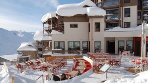 Un área de comedor al aire libre cubierta de nieve, con sillas y mesas a rayas rojas y blancas, rodeada por un edificio con múltiples balcones y ventanas.