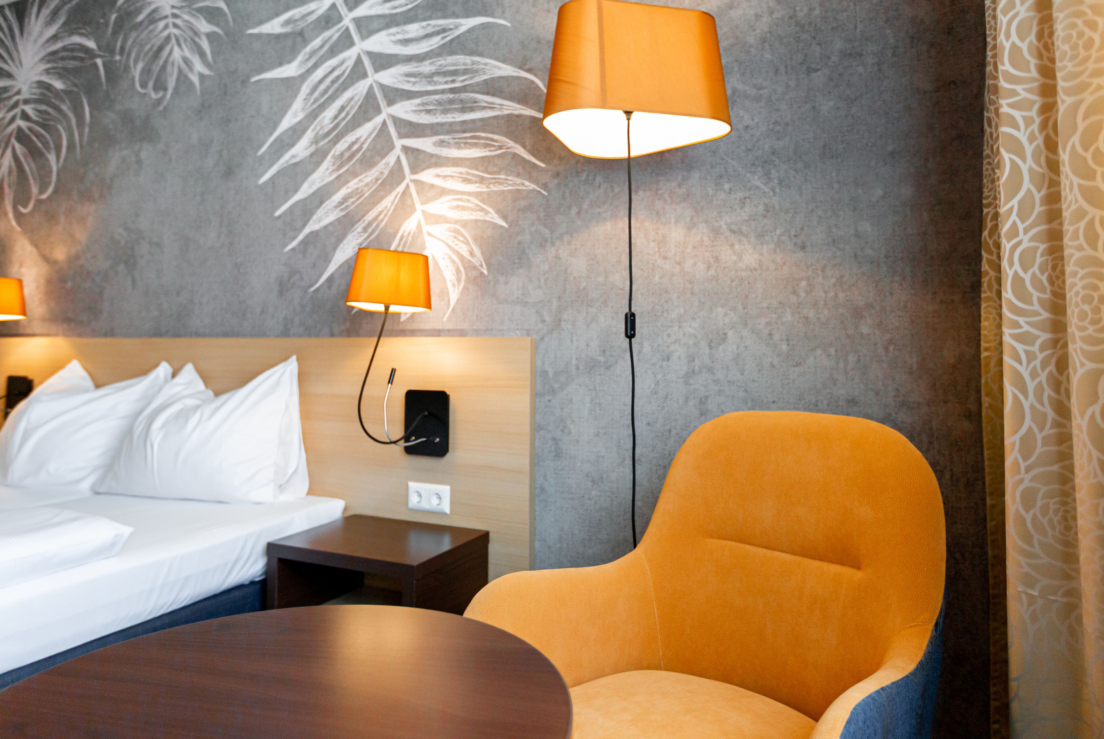 A hotel room with a bed, two bedside lamps, a wooden nightstand, a round table, and a large orange armchair.
