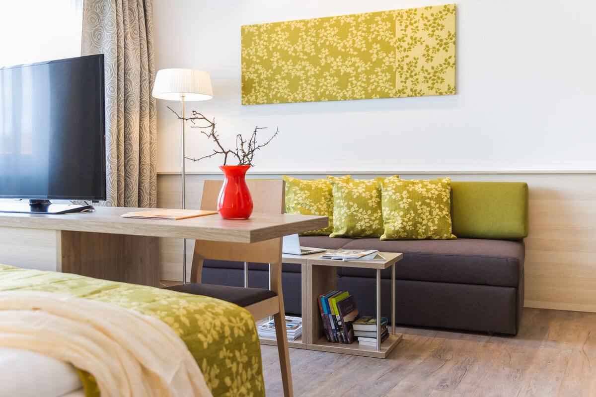 A modern hotel room with a flat-screen TV, a desk with a lamp, a red vase, a sofa with floral cushions, and a bookshelf.