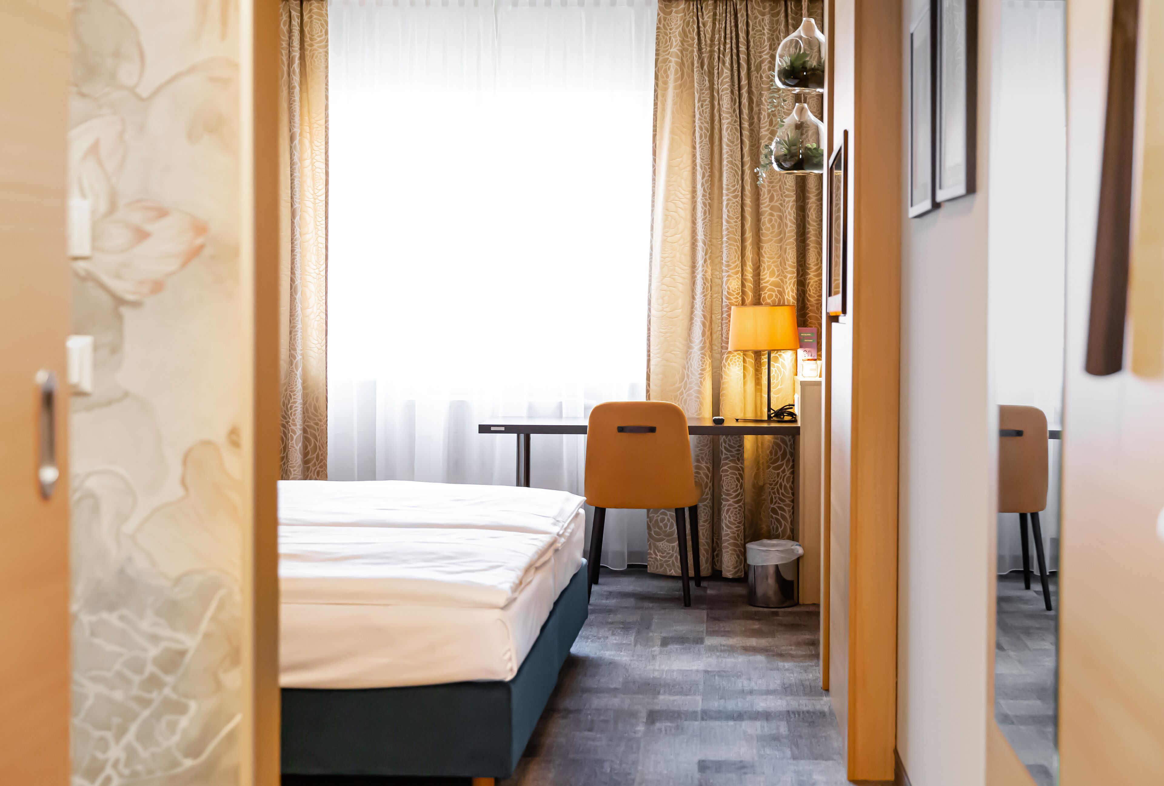 Quarto de hotel com cama, escrivaninha e cadeira. Há uma janela com cortinas, uma luminária na escrivaninha e um cesto de lixo.