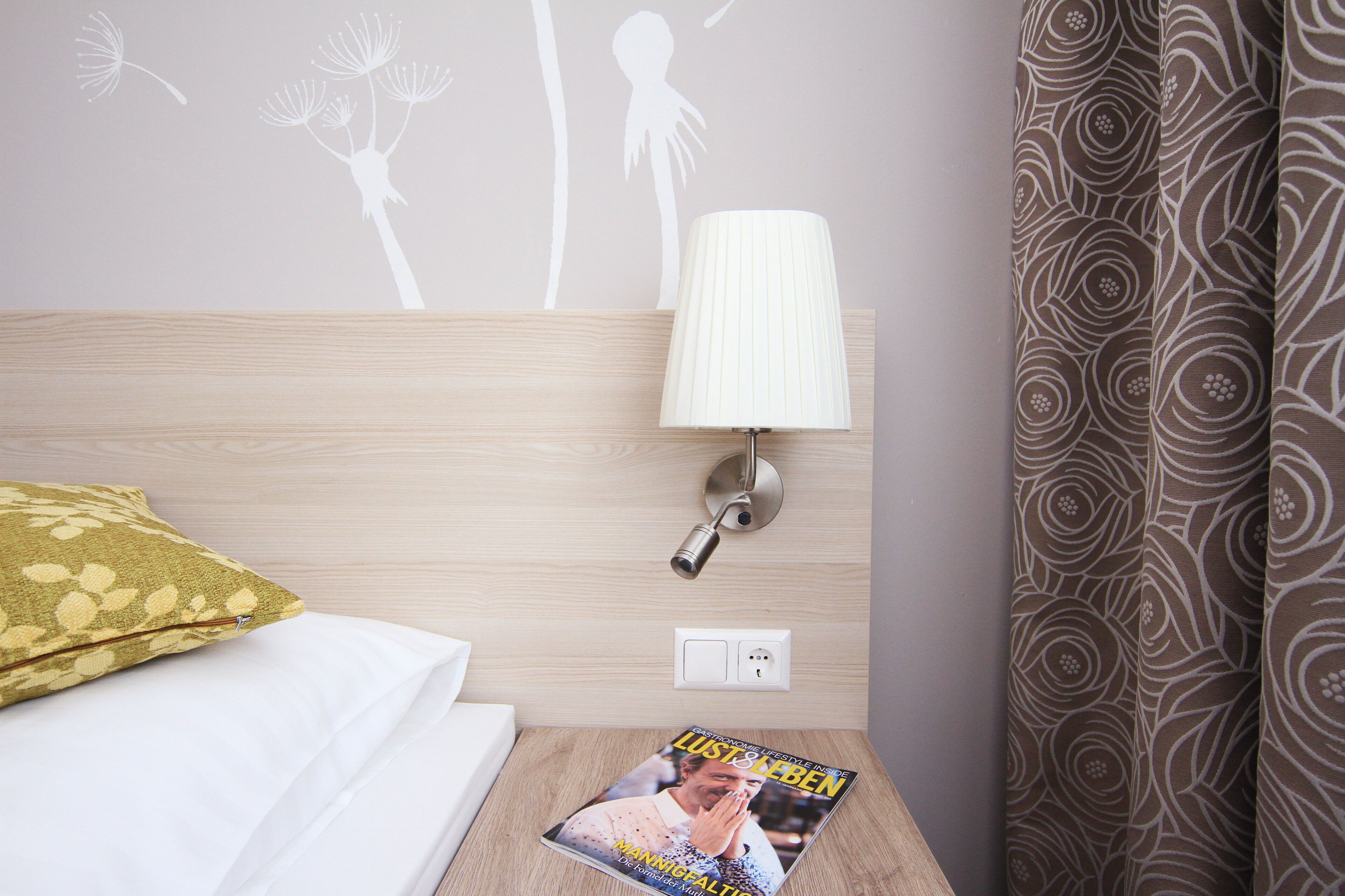 Une chambre d’hôtel avec un lit, une table de chevet avec une lampe, un magazine et un rideau à motifs.